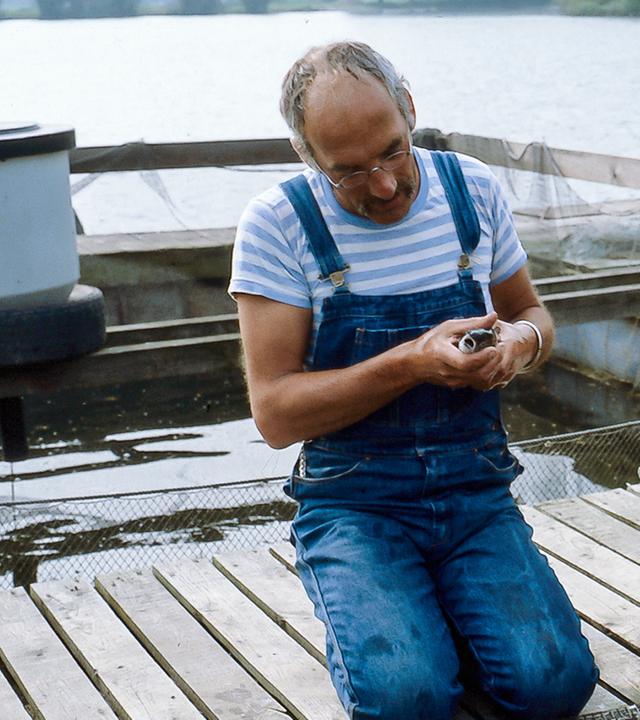 Peter sitzt auf einem Steg am Wasser, neben ihm ein Mann, der einen Kescher hält.