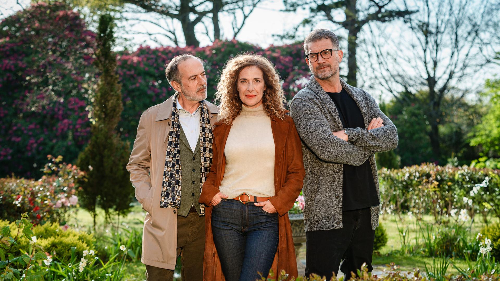 "Rosamunde Pilcher: Wo Liebe aufhört und wo sie beginnt": In einem Garten mit blühenden Pflanzen: Kate Evans (Clelia Sarto) steht in der Mitte zwischen Andrew Evans (Simon Böer) und Yamen Nicholls (Merab Ninidze)