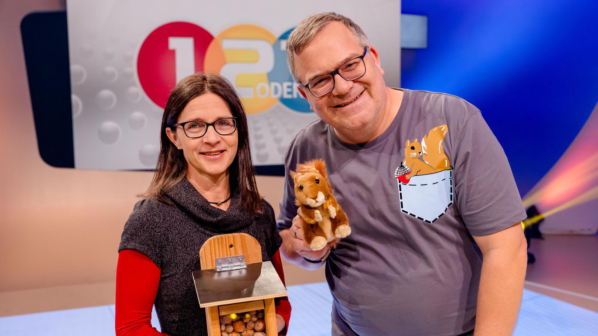 Eichhörnchen-Expertin Dr. Tine Meier steht im "1, 2 oder 3"-Studio und hält eine Futterstation mit Nüssen in den Händen. Elton steht daneben und hält ein Stoff-Eichhörnchen in der Hand.