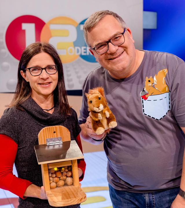 Eichhörnchen-Expertin Dr. Tine Meier steht im "1, 2 oder 3"-Studio und hält eine Futterstation mit Nüssen in den Händen. Elton steht daneben und hält ein Stoff-Eichhörnchen in der Hand.
