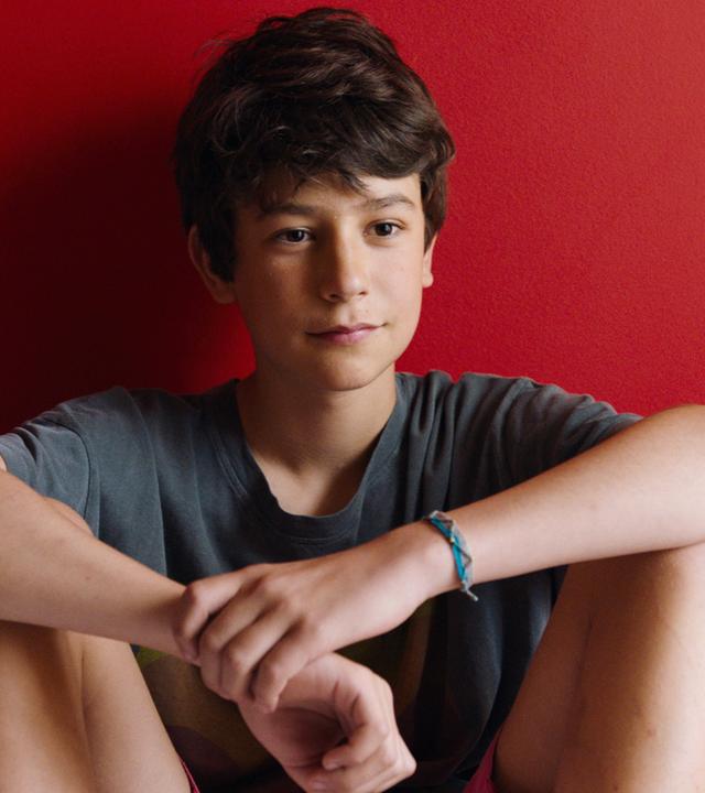 Der 13jährige Rémi sitzt mit angewinkelten Beinen auf dem Bett vor einer rot gestrichenen Wand.