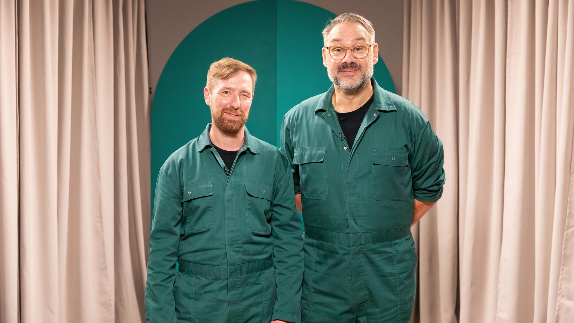 Zwei Menschen in grünen Overalls blicken in die Kamera