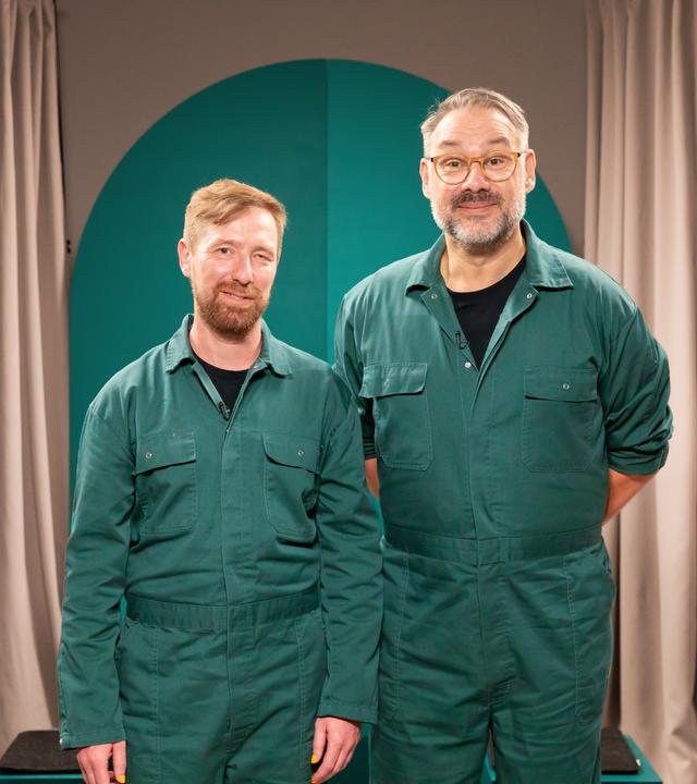 Zwei Menschen in grünen Overalls blicken in die Kamera