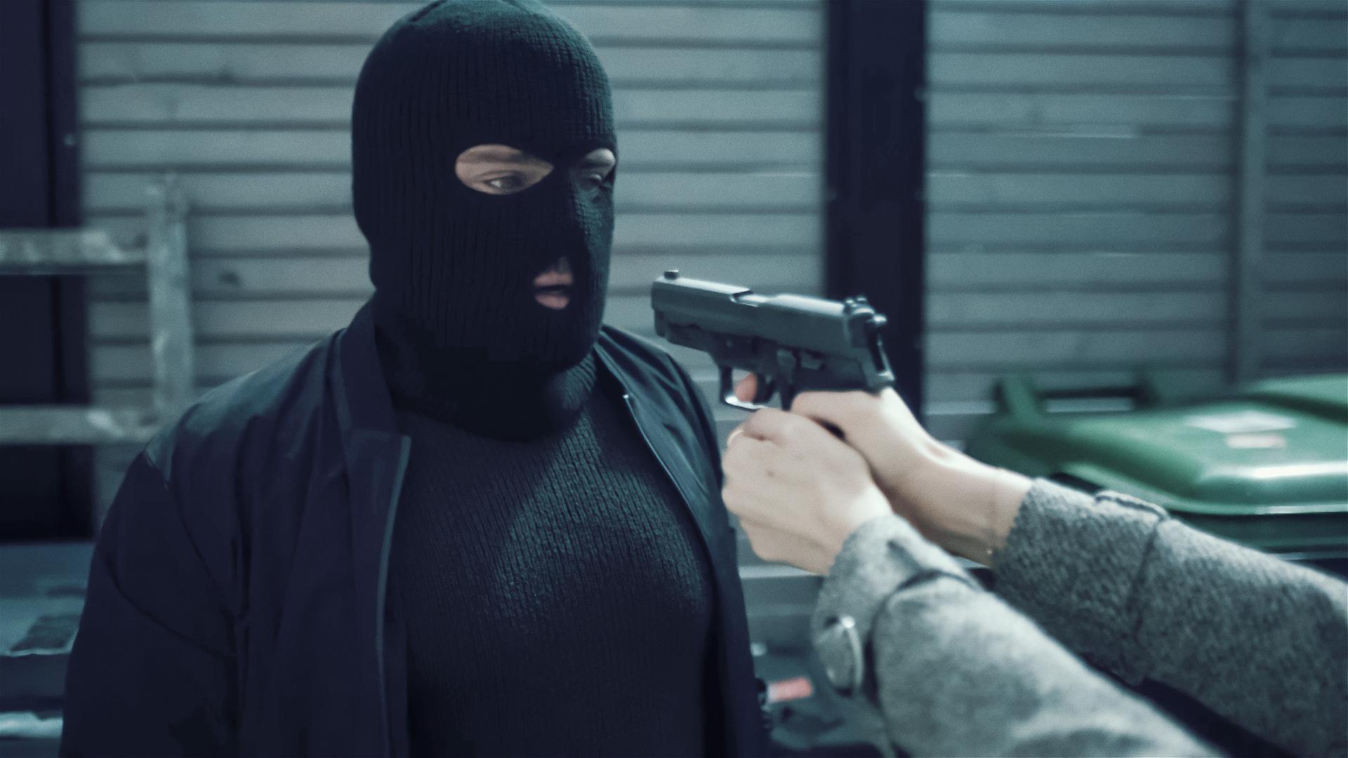 Ein Mann mit einer schwarzen Sturmmaske wird mit einer Pistole bedroht. Die Waffe liegt in den Händen einer Frau, von der man allerdings nur die Unterarme sehen kann.