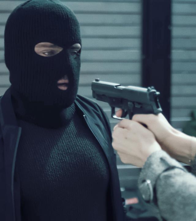 Ein Mann mit einer schwarzen Sturmmaske wird mit einer Pistole bedroht. Die Waffe liegt in den Händen einer Frau, von der man allerdings nur die Unterarme sehen kann.