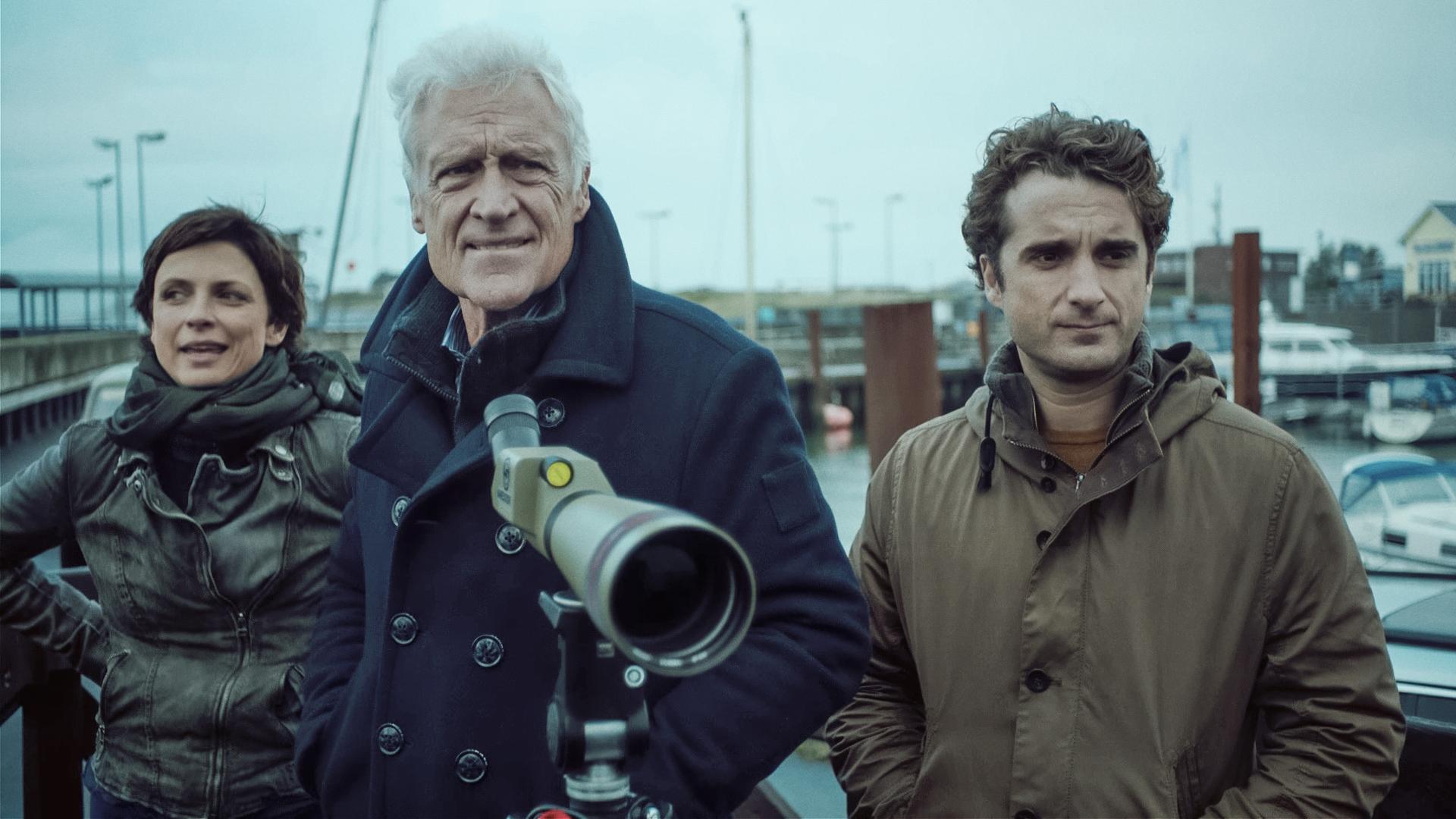 Behrendsen, Clüver und Feldmann (Julia Brendler, Robert Atzorn, Oliver Wnuk), alle in Wintermänteln gekleidet, stehen mit einem Fernglas, das auf einem Stativ befestigt sind, im Hafen. 