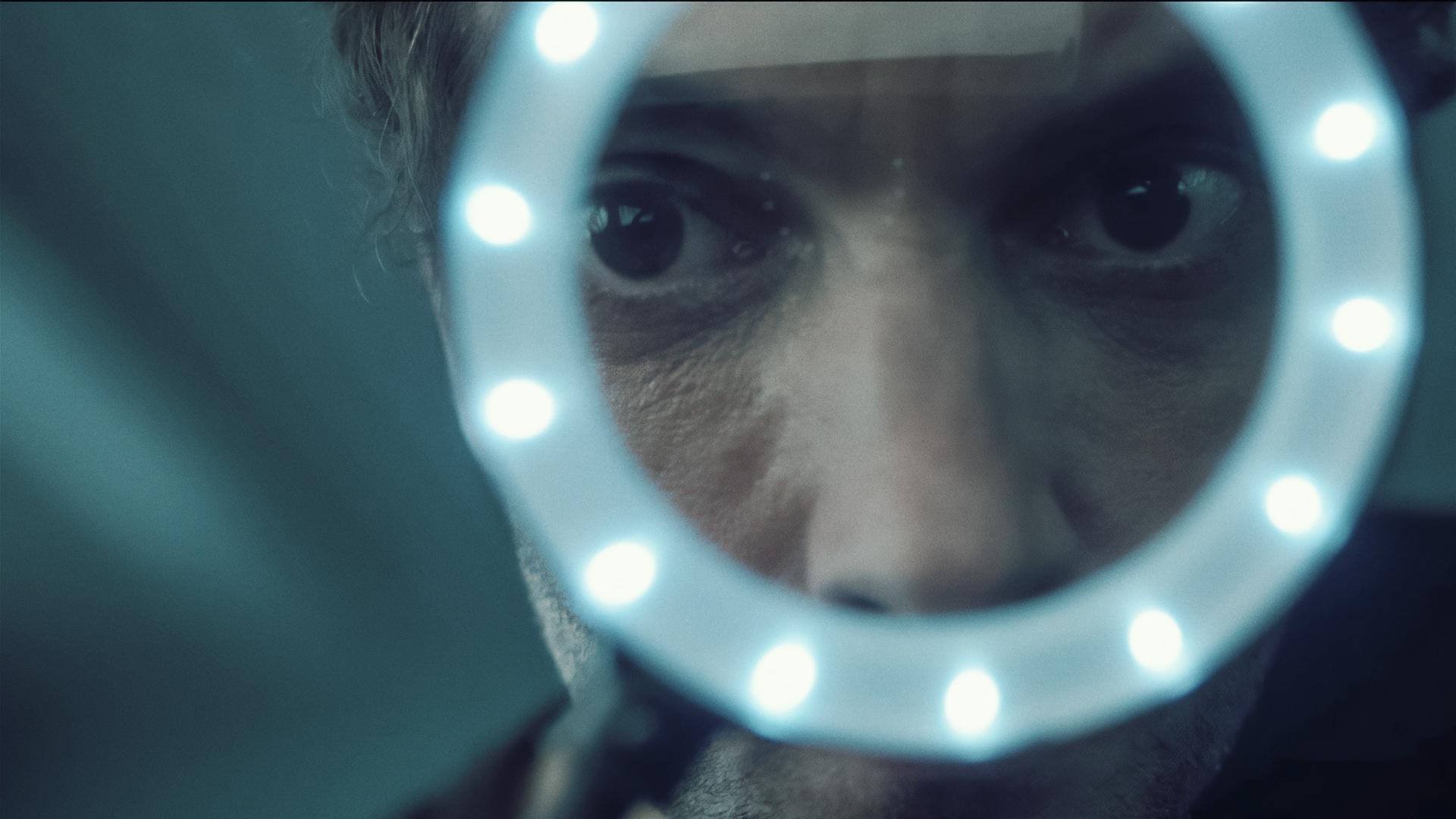 Aus der Untersicht blickt man zu Feldmann (Oliver Wnuk) auf, der durch ein beleuchtetes Vergrößerungsglas schaut. Durch die Lupe ist sein Gesicht verzerrt, seine dunkel unterlaufenen Augen erscheinen riesig. 