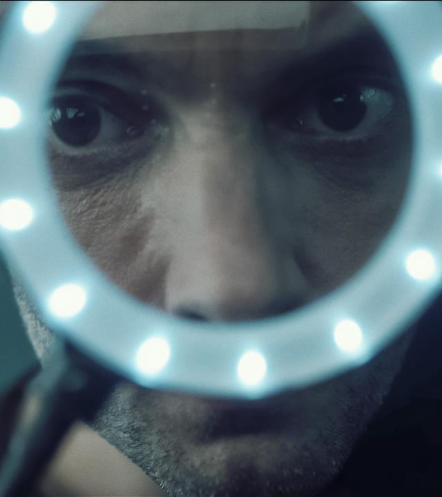 Aus der Untersicht blickt man zu Feldmann (Oliver Wnuk) auf, der durch ein beleuchtetes Vergrößerungsglas schaut. Durch die Lupe ist sein Gesicht verzerrt, seine dunkel unterlaufenen Augen erscheinen riesig. 