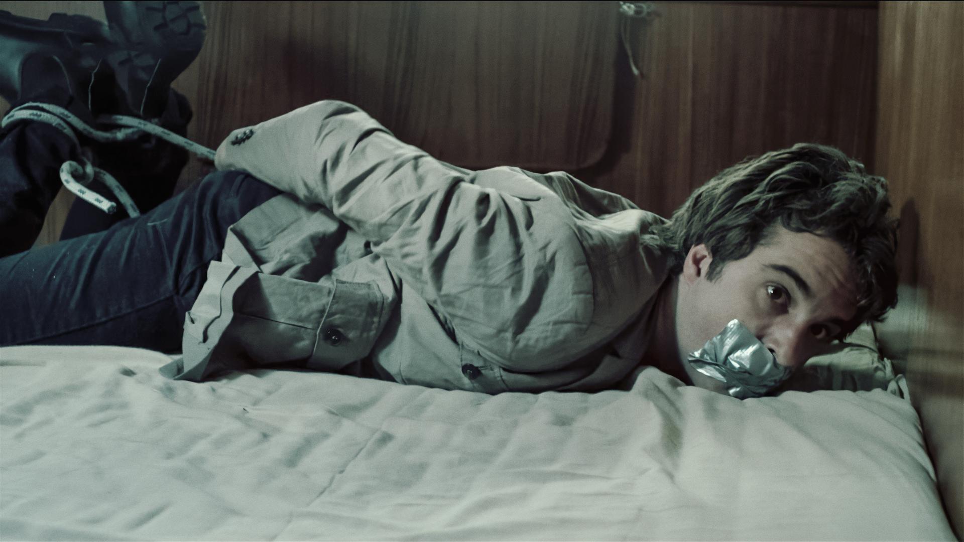 Kommissar Hinnerk Feldmann (Oliver Wnuk) liegt gefesselt und geknebelt bäuchlings auf einem Bett. 