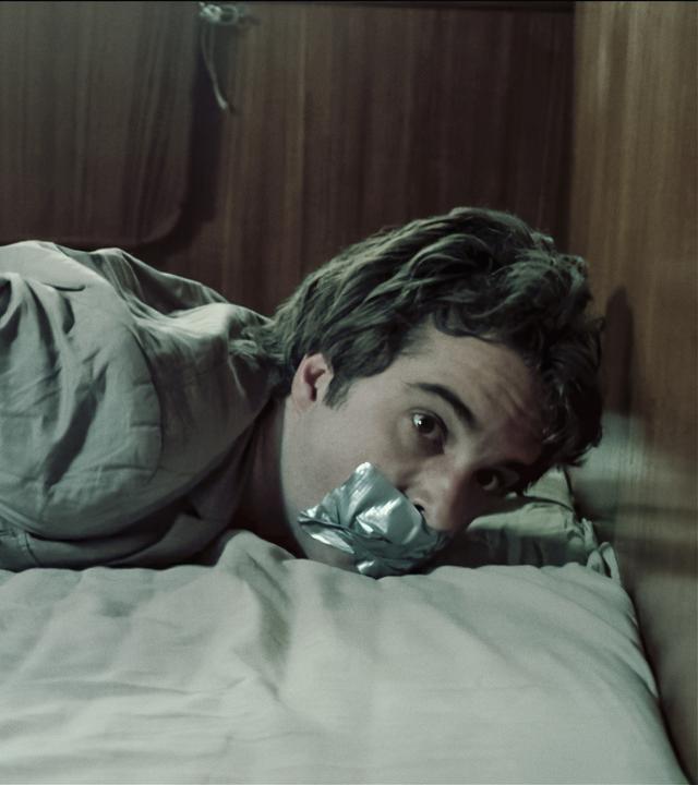 Kommissar Hinnerk Feldmann (Oliver Wnuk) liegt gefesselt und geknebelt bäuchlings auf einem Bett. 