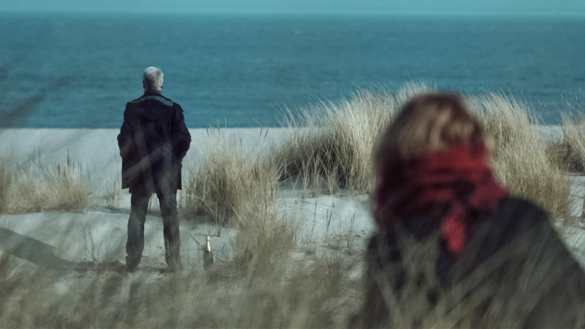 Clüver steht auf einer Düne und blickt auf das blaue Meer vor ihm. Man kann ihn nur von hinten sehen. Zu seinen Füßen steht eine Sektflasche. Am rechten unteren Bildrand nähert sich eine blonde Frau, die ebenfalls nur von hinten zu sehen ist. 