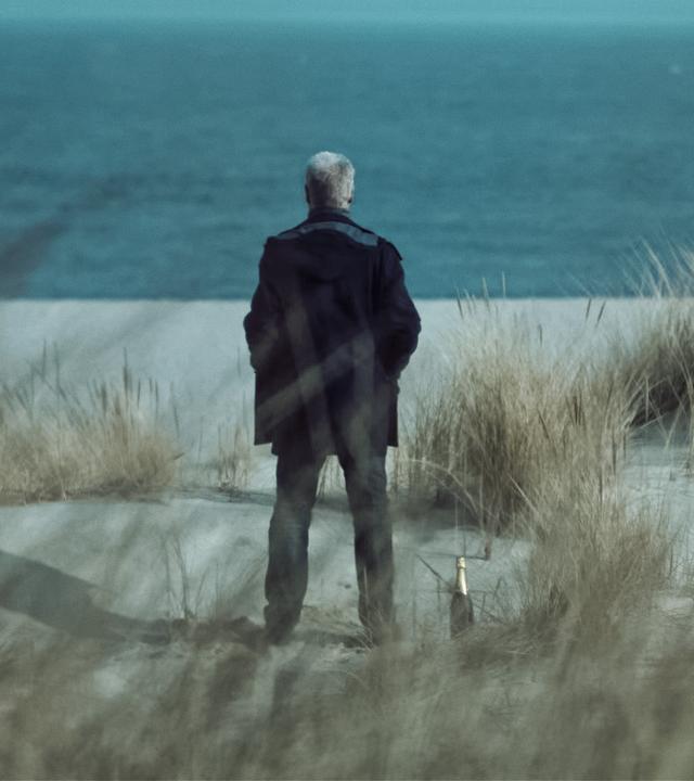 Clüver steht auf einer Düne und blickt auf das blaue Meer vor ihm. Man kann ihn nur von hinten sehen. Zu seinen Füßen steht eine Sektflasche. Am rechten unteren Bildrand nähert sich eine blonde Frau, die ebenfalls nur von hinten zu sehen ist. 