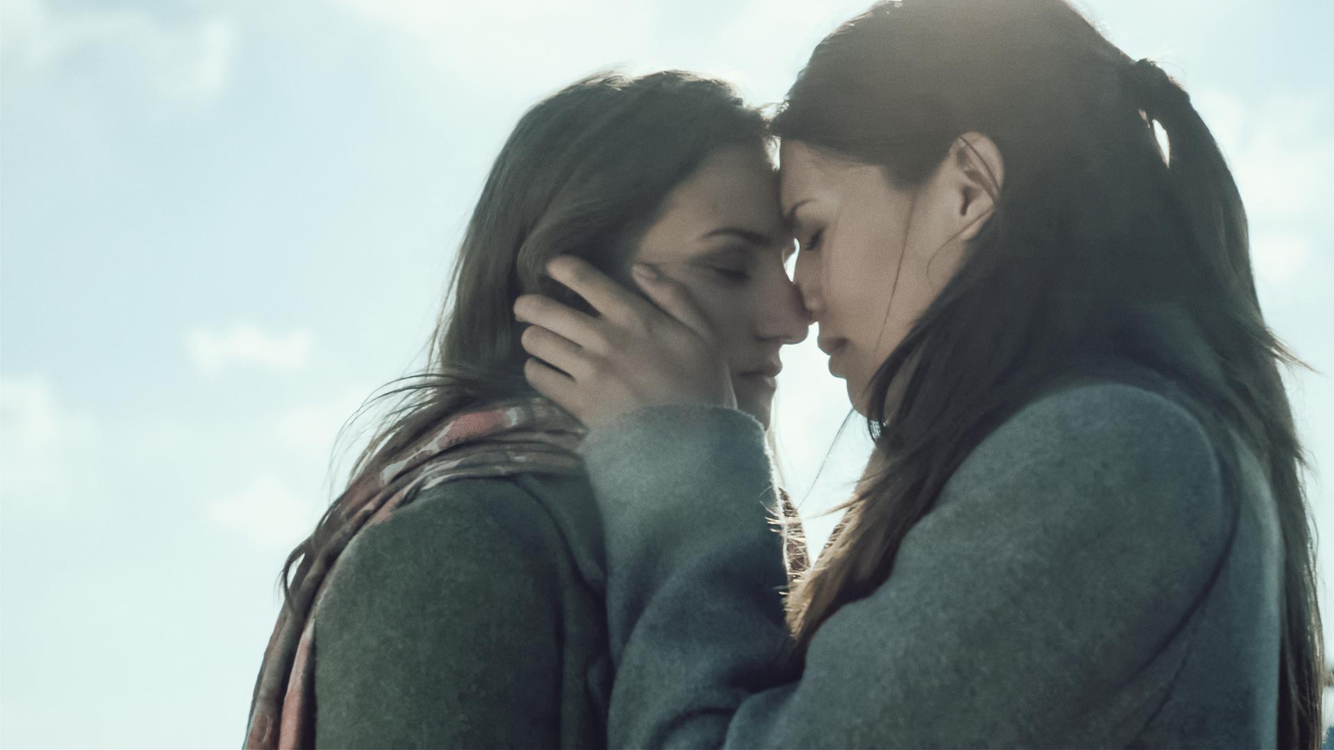 Zwei junge Frauen im Freien: Die beiden haben die Köpfe aneinander gelegt, eine hält die Wange der anderen in einer romantischen Geste kurz vor einem Kuss. 