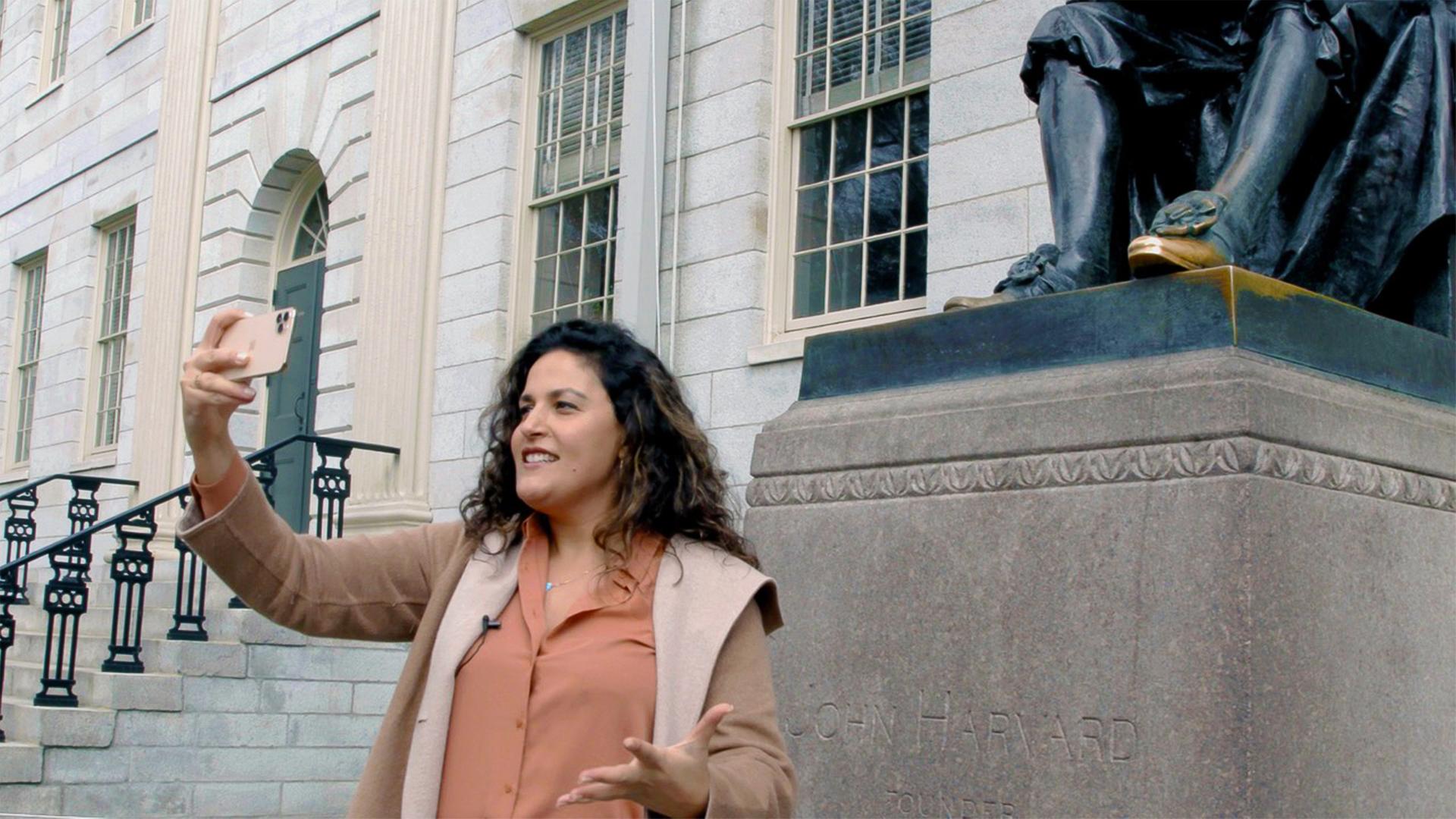 Eine Frau steht vor einem großen kassizistischen Gebäude mit einer Statue vor dem Eingang und macht ein Selfie.