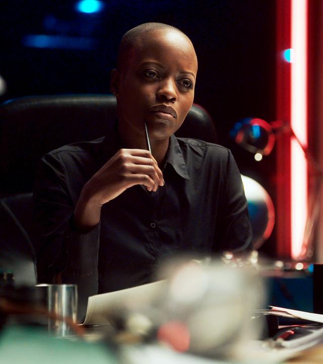 Florence Kasumba sitzt an einem überfüllten Schreibtisch, hält sich nachdenklich einen Stift vors Gesicht und schaut an der Kamera vorbei