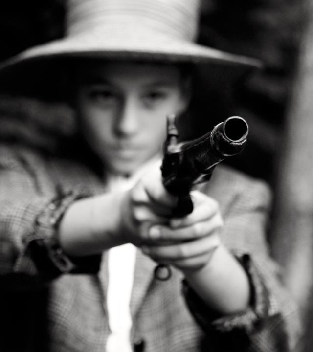 Nachgestellte Szene. Unscharfes Schwarz-Weiß-Foto: Ein Kind mit Hut hält einen altmodischen Revolver in beiden Händen.