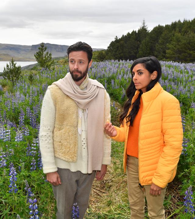 "Das Traumschiff: Island": Auf einem mit Lupinen bewachsenen Hügel: Dr. Jessica Delgado (Collien Fernandes) steht neben Jon (Pétur Óskar). Sie blicken erwartungsvoll seitlich nach vorne. Im Hintergrund befindet sich ein Tannenwald, hinter diesem liegt das Meer.