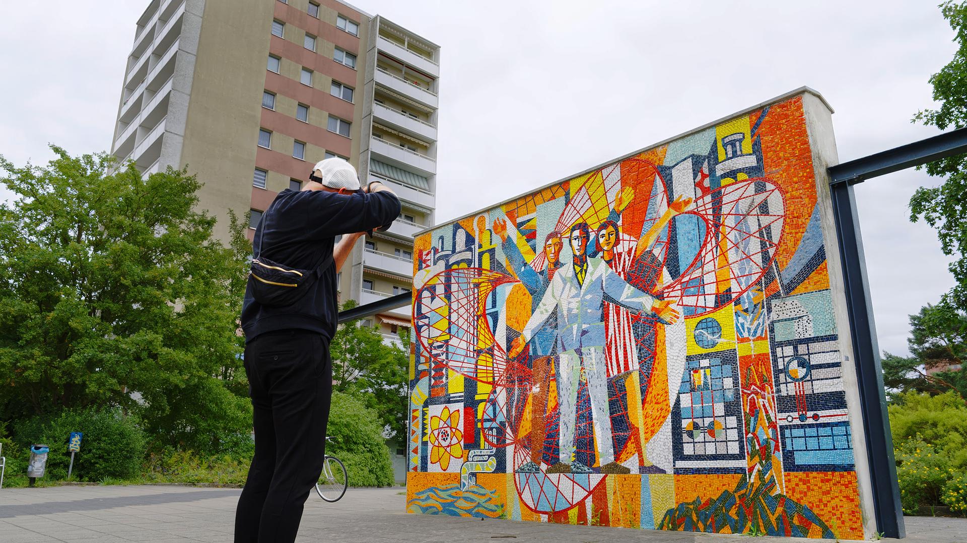 Das Bild zeigt einen Fotografen, der ein farbenfrohes Mosaikkunstwerk vor einer Reihe von Plattenbauten in Eisenhüttenstadt ablichtet. Der Fotograf ist von hinten zu sehen, gekleidet in schwarzer Kleidung und einer weißen Baseballkappe. Er hält eine Kamera vor das Gesicht und richtet den Fokus auf ein Kunstwerk. Die Szene wird von den grauen und beigen Hochhäusern im Hintergrund eingerahmt, Wohnblöcke im Stil der DDR-Plattenbauweise. 