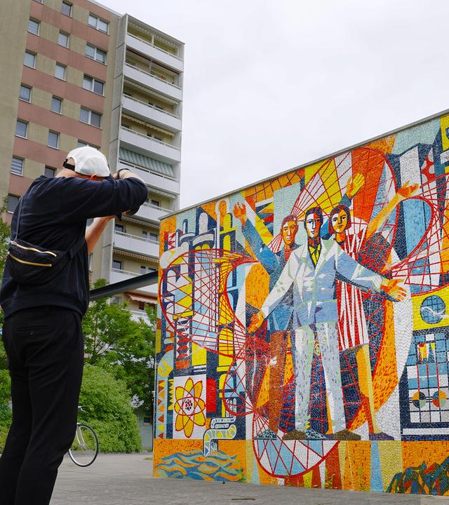 Das Bild zeigt einen Fotografen, der ein farbenfrohes Mosaikkunstwerk vor einer Reihe von Plattenbauten in Eisenhüttenstadt ablichtet. Der Fotograf ist von hinten zu sehen, gekleidet in schwarzer Kleidung und einer weißen Baseballkappe. Er hält eine Kamera vor das Gesicht und richtet den Fokus auf ein Kunstwerk. Die Szene wird von den grauen und beigen Hochhäusern im Hintergrund eingerahmt, Wohnblöcke im Stil der DDR-Plattenbauweise. 