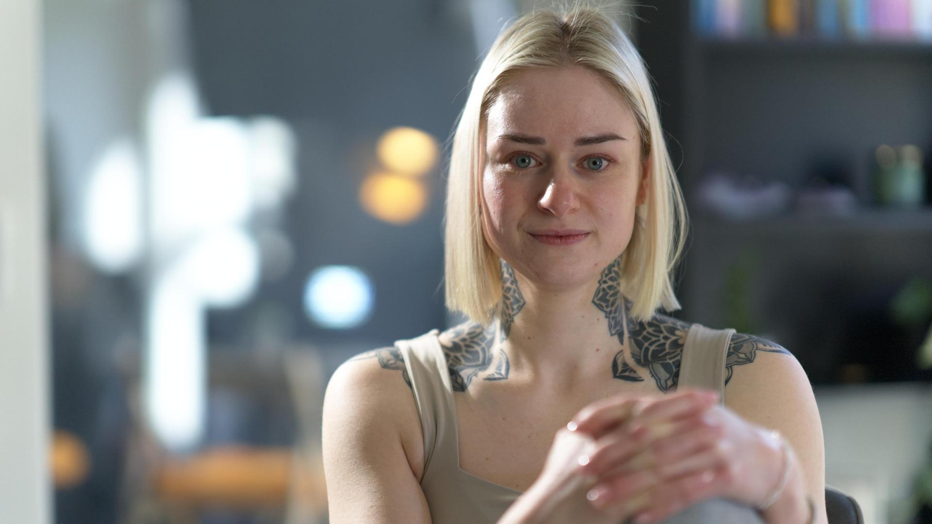 Sandrine hat kinnlange blonde Haare und ein großes Tattoo auf den Schultern und am Hals. Sie schaut direkt in die Kamera und legt die Hände auf ihr angezogenes Knie.