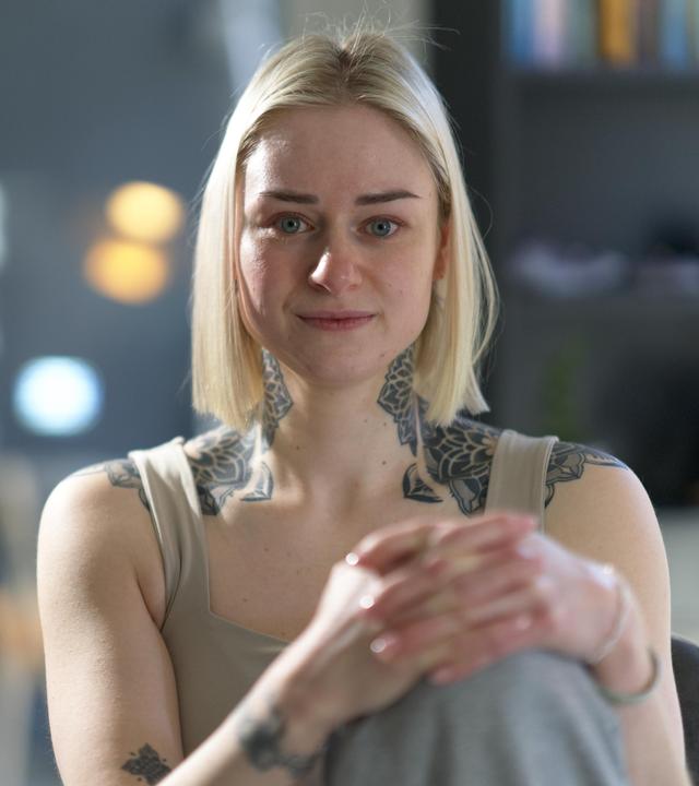 Sandrine hat kinnlange blonde Haare und ein großes Tattoo auf den Schultern und am Hals. Sie schaut direkt in die Kamera und legt die Hände auf ihr angezogenes Knie.