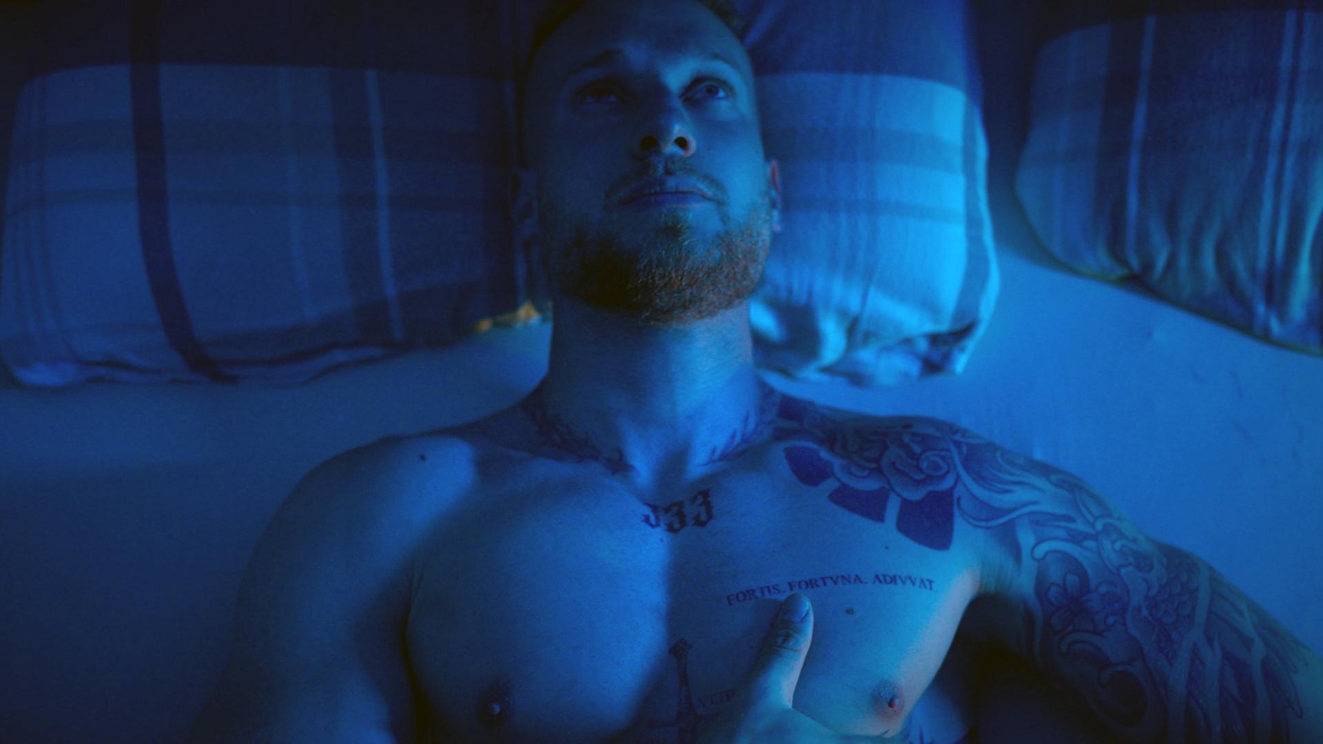 Tom sitzt oberkörperfrei auf seinem Bett. Der Raum ist in ein blaues Licht getaucht. 