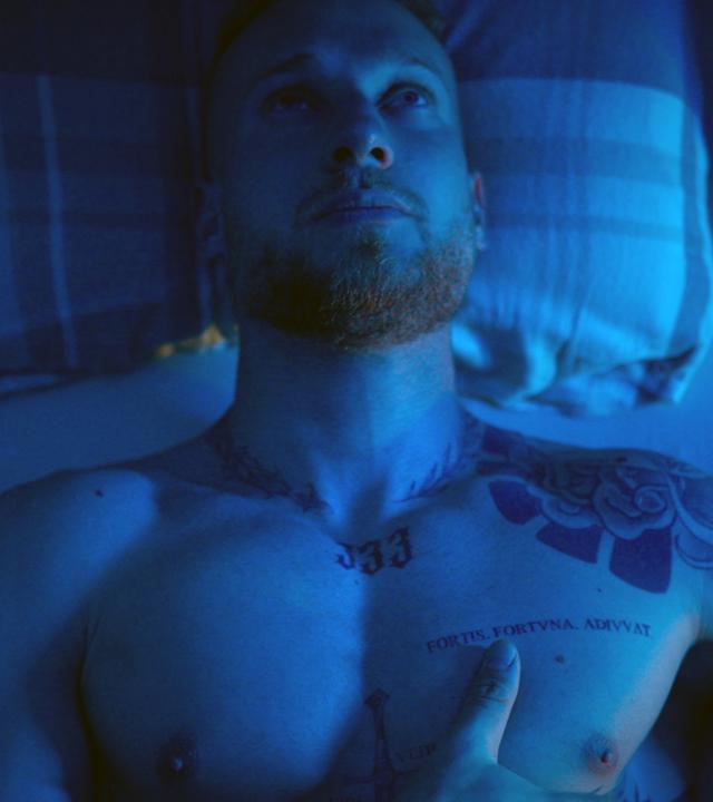 Tom sitzt oberkörperfrei auf seinem Bett. Der Raum ist in ein blaues Licht getaucht. 
