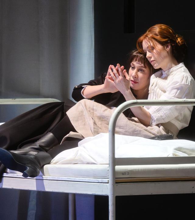 Zwei Frauen liegen in einem Bett auf der Bühne und berühren sich mit einer Hand.