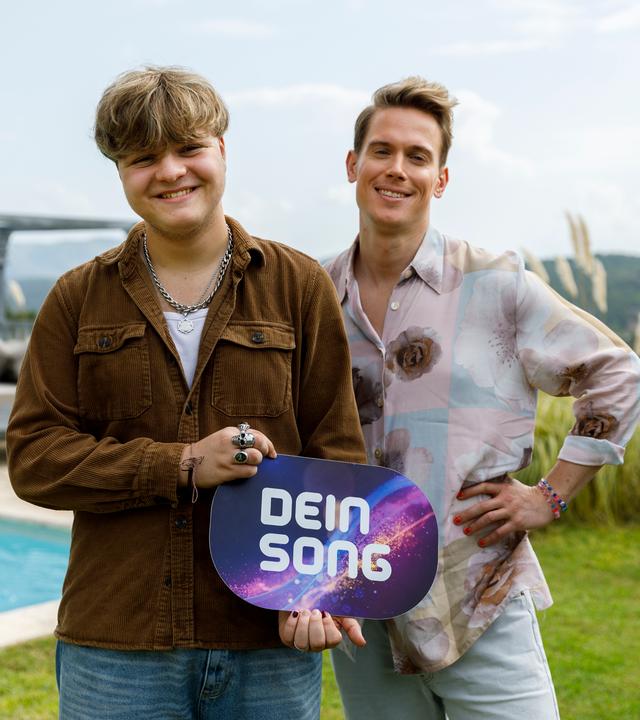 Felix und Conny stehen nebeneinander. Felix hält ein Dein Song-Schild in den Händen, CONNY hat seinen rechten Arm in die Hüften gestemmt. Beide lächeln.