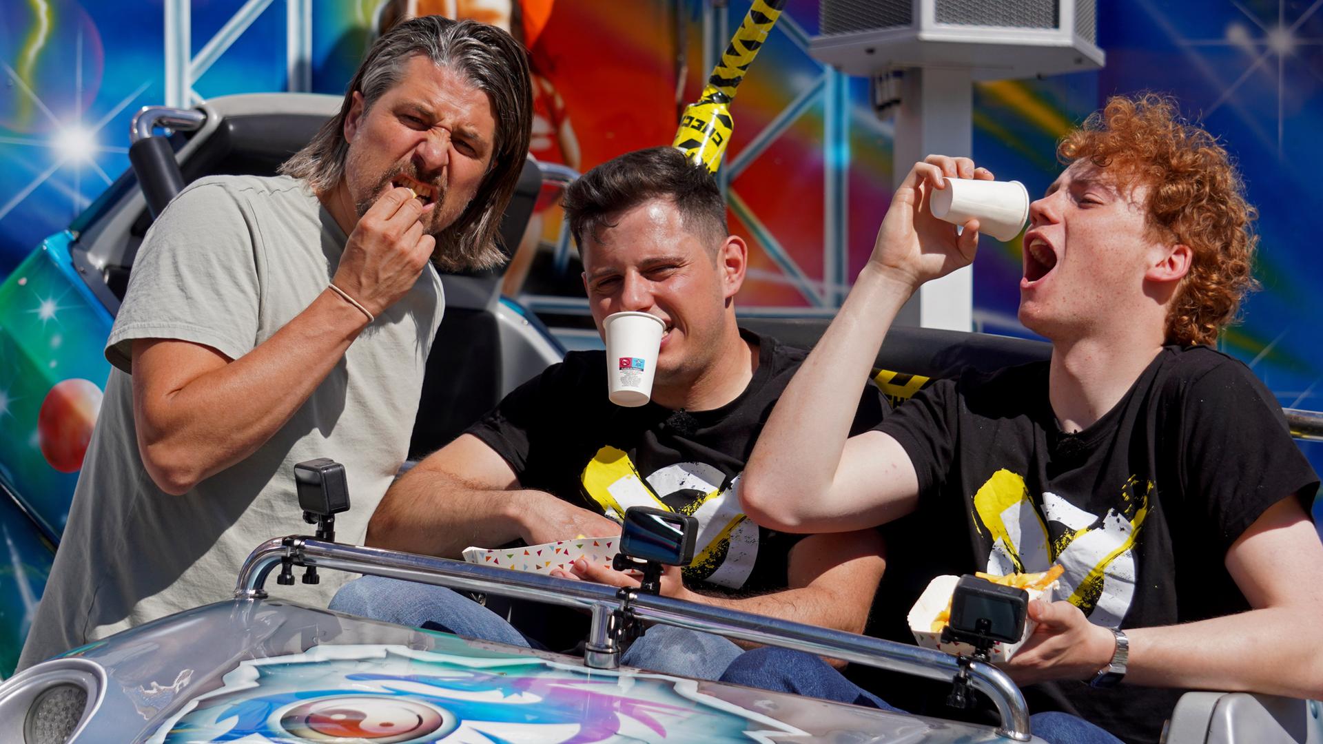 Checkpoint: Tommy Scheel lehnt sich an einen Wagen eines Jahrmarkt-Fahrgeschäfts, in dem Cem und Jona sitzen und so tun, als ob sie aus Bechern trinken.