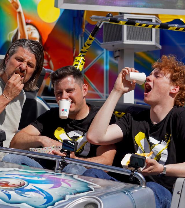 Checkpoint: Tommy Scheel lehnt sich an einen Wagen eines Jahrmarkt-Fahrgeschäfts, in dem Cem und Jona sitzen und so tun, als ob sie aus Bechern trinken.