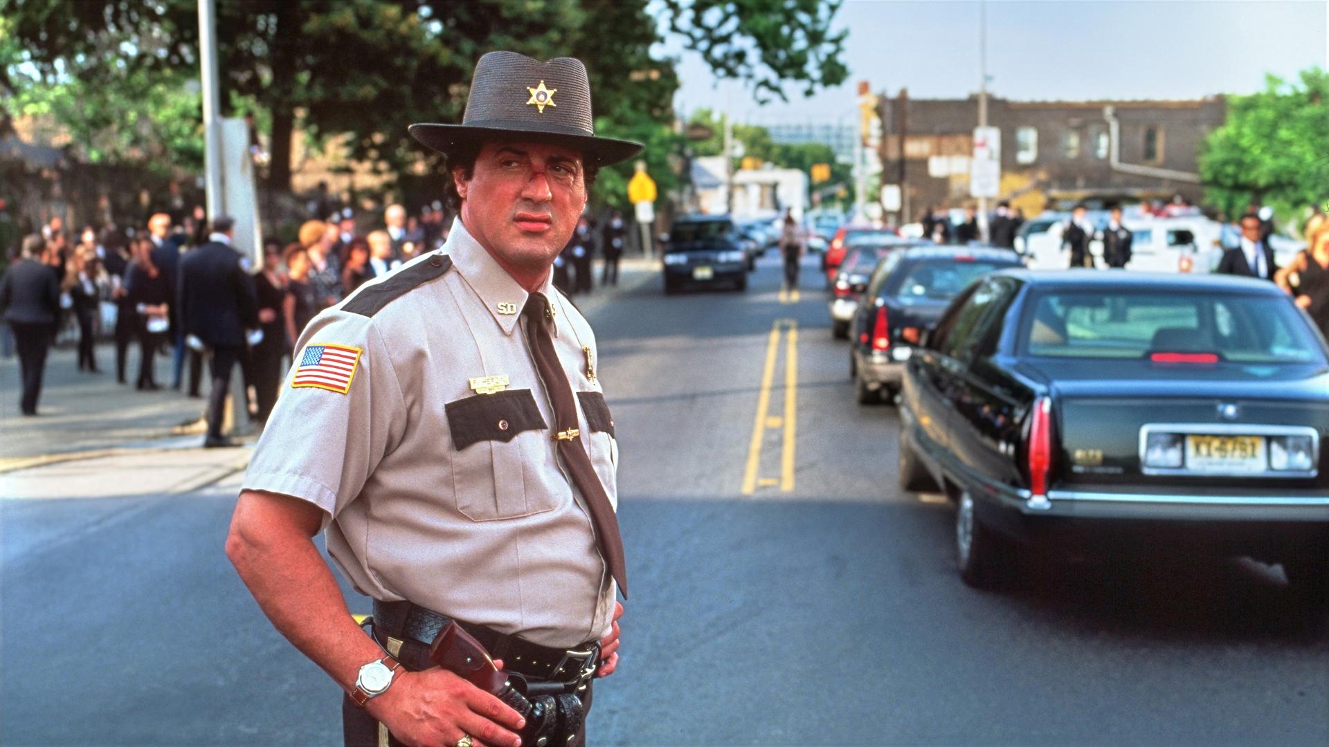 Sheriff Freddy Heflin steht in Dienstkleidung auf einer Straße und blickt zur Seite. Im Hintergrund fahren mehrere Autos, während Menschen am Straßenrand stehen.