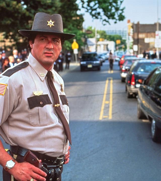 Sheriff Freddy Heflin steht in Dienstkleidung auf einer Straße und blickt zur Seite. Im Hintergrund fahren mehrere Autos, während Menschen am Straßenrand stehen.