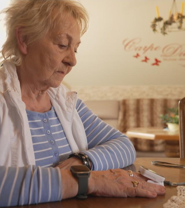Eine Frau sitzt am Tisch und blickt auf das Sauerstoffmessgerät an ihrem Finger.
