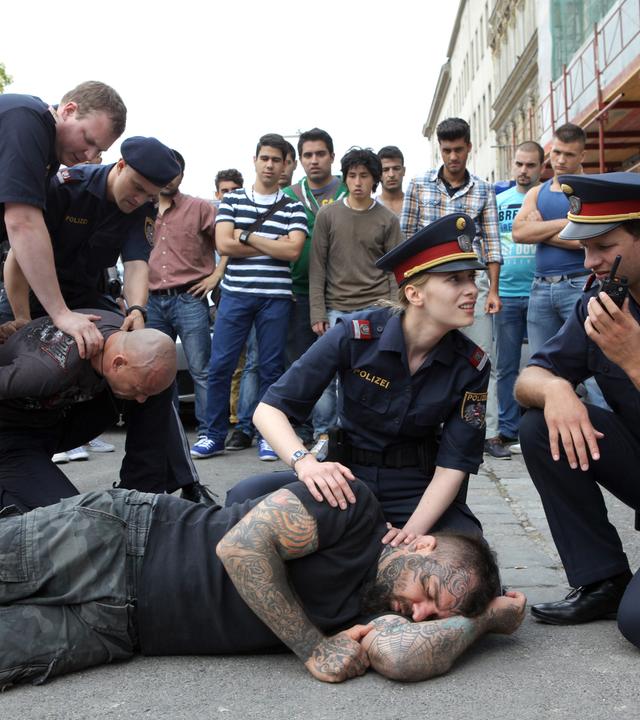 Das Bild zeigt eine Szenen aus dem Film „CopStories“. Es spielt auf einer Straße, wo eine Gruppe von Polizisten und Zivilisten zu sehen ist.   Im Vordergrund liegen zwei Männer auf dem Boden. Einer ist tätowiert, trägt ein schwarzes T-Shirt und sieht bewusstlos aus. Der andere Mann, der am Boden kniet, ist ein Polizist, der ihn festhält. Zwei weitere Polizeibeamte versuchen ebenfalls, die Situation zu kontrollieren. Die Polizistin mit blonden Haaren und einer Polizeimütze kniet neben dem tätowierten Mann und kümmert sich um ihn, während ein anderer Polizist anspricht oder möglicherweise Funksprüche abgibt.  Im Hintergrund stehen mehrere Zivilisten in unterschiedlichen Kleidungsstilen und beobachten die Situation. Einige scheinen interessiert und möglicherweise besorgt zu sein. Hinter ihnen ist ein Polizeiauto mit sichtbaren Lizenznummern geparkt. Es sind auch Baustrukturen und Bäume in der Umgebung sichtbar, die der Szene ein städtisches Ambiente verleihen.  Die gesamte Szene vermittelt einen Eindruck von Chaos und Spannung, da die Polizisten versuchen, eine potenziell gefährliche Situation unter Kontrolle zu bringen.