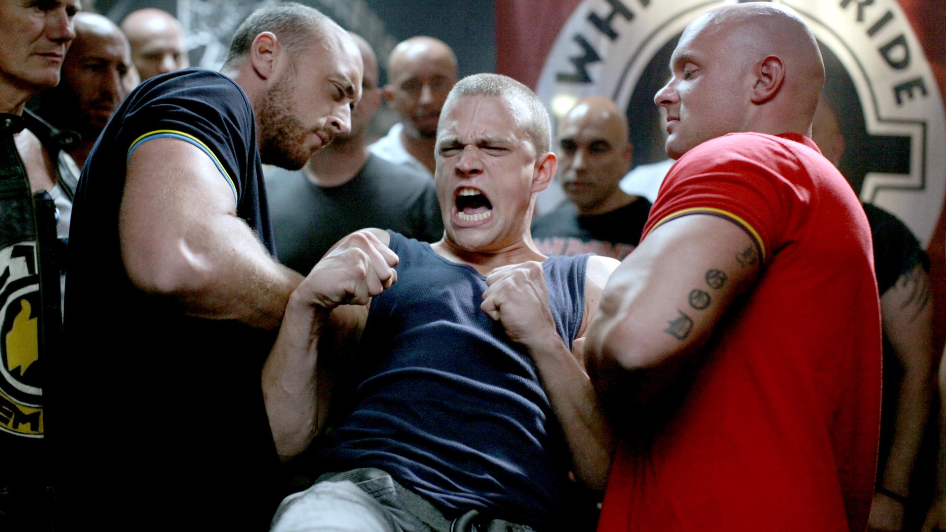 Das Bild zeigt eine Szene aus dem Film "CopStories – Bist du deppert". In einem dunklen und angespannten Raum stehen mehrere muskulöse Männer, die intensiv in eine Auseinandersetzung verwickelt sind.   Im Vordergrund sieht man einen jungen Mann mit kurzgeschorenen Haaren, der ein ärmelloses Oberteil trägt. Sein Gesicht ist verzerrt vor Wut oder Anstrengung, während er mit beiden Fäusten in der Luft kämpferisch wirkt. Er wird von zwei kräftigen Männern festgehalten, einer von ihnen trägt ein dunkelblaues Shirt mit gelben Akzenten, der andere ein rotes Shirt und hat sichtbare Tätowierungen auf seinem rechten Arm.  Im Hintergrund sind weitere Männer zu sehen, die aufmerksam auf die Szene reagieren; sie sind nahezu alle glatzköpfig oder haben sehr kurze Haare. Der Raum hat eine düstere Atmosphäre, mit wenig Licht und einer Wand, die mit dem Schriftzug "WHITE PRIDE" dekoriert ist.   Die gesamte Komposition des Bildes vermittelt ein Gefühl von Aggression und Konflikt, und die Körperhaltungen der Figuren zeigen, dass sich kurz vor einem entscheidenden Moment etwas anbahnt.