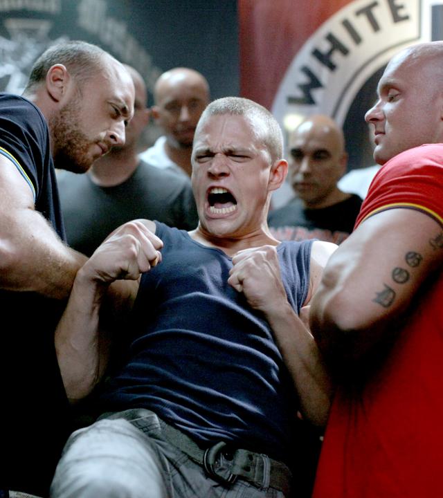Das Bild zeigt eine Szene aus dem Film "CopStories – Bist du deppert". In einem dunklen und angespannten Raum stehen mehrere muskulöse Männer, die intensiv in eine Auseinandersetzung verwickelt sind.   Im Vordergrund sieht man einen jungen Mann mit kurzgeschorenen Haaren, der ein ärmelloses Oberteil trägt. Sein Gesicht ist verzerrt vor Wut oder Anstrengung, während er mit beiden Fäusten in der Luft kämpferisch wirkt. Er wird von zwei kräftigen Männern festgehalten, einer von ihnen trägt ein dunkelblaues Shirt mit gelben Akzenten, der andere ein rotes Shirt und hat sichtbare Tätowierungen auf seinem rechten Arm.  Im Hintergrund sind weitere Männer zu sehen, die aufmerksam auf die Szene reagieren; sie sind nahezu alle glatzköpfig oder haben sehr kurze Haare. Der Raum hat eine düstere Atmosphäre, mit wenig Licht und einer Wand, die mit dem Schriftzug "WHITE PRIDE" dekoriert ist.   Die gesamte Komposition des Bildes vermittelt ein Gefühl von Aggression und Konflikt, und die Körperhaltungen der Figuren zeigen, dass sich kurz vor einem entscheidenden Moment etwas anbahnt.