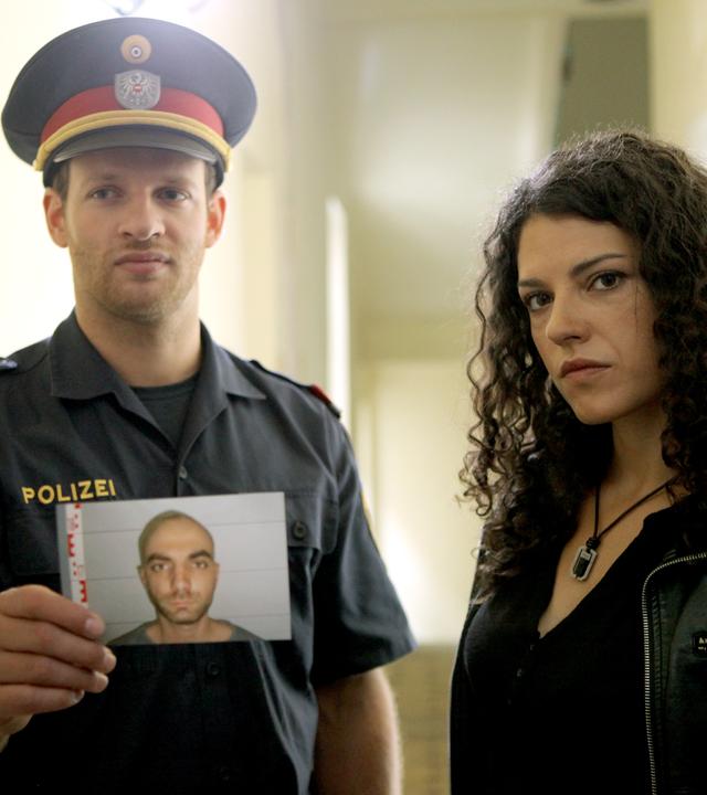 Das Bild zeigt zwei Personen in einem Innenraum. Auf der linken Seite steht ein Mann in einer Polizeiuniform mit einem schwarzen Shirt und einer Polizeijacke. Er trägt eine Kappe mit dem Wappen der Polizei und hält ein Foto in der Hand, das eine junge männliche Person zeigt, die ernst schaut. Der Mann hat einen neutralen Gesichtsausdruck.  Rechts von ihm steht eine Frau mit lockigem, dunklem Haar. Sie trägt eine schwarze Lederjacke und hat einen ernsten Gesichtsausdruck. Ihr Blick ist direkt auf die Kamera gerichtet. Der Hintergrund des Bildes zeigt einen langen Flur mit Wänden in hellem Farbton, der von einer Deckenbeleuchtung erhellt wird.  Die Szene wirkt angespannt, was durch die Körperhaltung der beiden Personen und ihre Ausdrücke verstärkt wird.