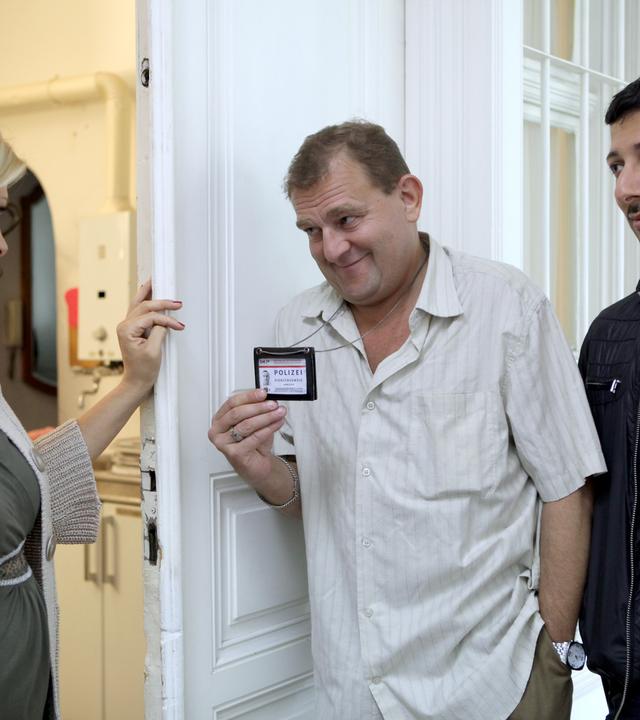 Auf dem Bild sind drei Personen in einem Innenraum zu sehen. Die Szene scheint in einer Wohnung zu spielen. An der linken Seite steht eine Frau mit blonden, welligen Haaren, die ein graues, figurbetontes Kleid trägt und eine Strickjacke anhat. Sie lächelt und interagiert mit den anderen Personen.  In der Mitte steht ein Mann, der ein khaki-farbenes Hemd trägt, mit einem schelmischen Lächeln auf dem Gesicht. Er hält eine Polizeimarke oder einen Ausweis in der Hand, der an einer Kette um seinen Hals hängt. Rechts neben ihm steht ein weiterer Mann mit kurzen, dunklen Haaren, der eine schwarze Jacke und ein T-Shirt trägt. Er sieht nachdenklich aus und schaut in Richtung des Mannes in der Mitte.  Im Hintergrund sind Wände in hellen Farben sowie eine Kücheneinheit zu erkennen. Die Türöffnung lässt darauf schließen, dass der Raum gut beleuchtet ist. Die Atmosphäre scheint freundlich, jedoch auch ein wenig angespannt, angesichts der Thematik, die im Kontext des Films behandelt wird.