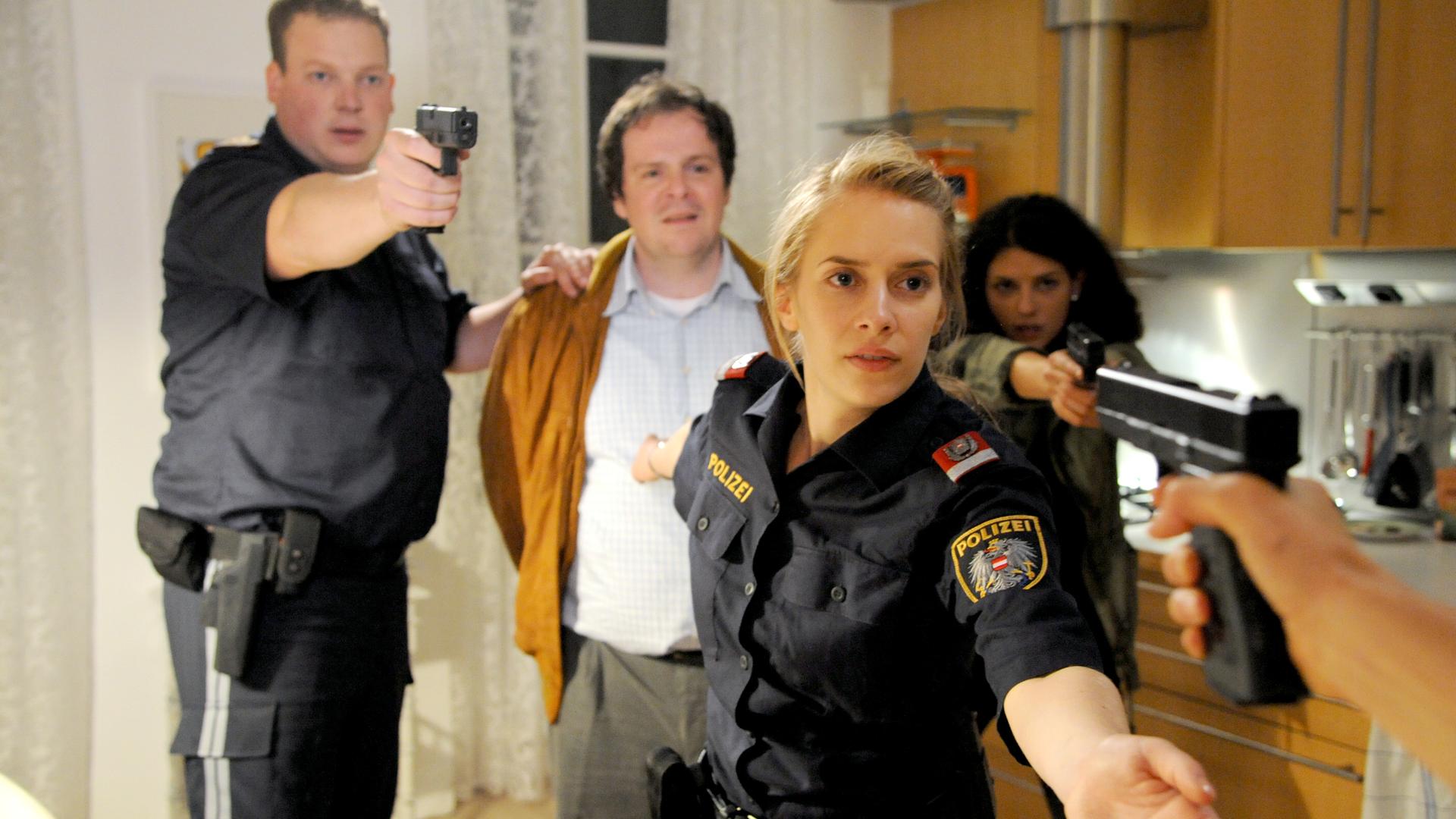 Das Bild zeigt eine spannungsgeladene Szene in einem Innenraum, vermutlich einer Wohnung oder einem Büro. Im Vordergrund stehen vier Personen. Eine Frau, gekleidet in eine Polizeiuniform, hält eine Waffe in der Hand und schaut entschlossen. Neben ihr steht ein Mann in einem hellen Hemd, der mit einem orangenen Mantel bedeckt ist; er hat eine besorgte Miene. Hinter ihm befinden sich zwei weitere Personen: eine Frau in einem olivfarbenen Outfit, die ebenfalls eine Waffe hält, und ein Mann in einer Polizeiuniform, der eine Waffe in Richtung des Betrachters richtet.  Der Raum hat helle Wände und eine moderne Holz-Küche im Hintergrund, die einige Küchengeräte und Ablagen zeigt. Vor den Fenstern befinden sich helle Vorhänge, die Licht hereinlassen. Die Pose der Charaktere deutet auf eine kritische Situation hin, in der sich alle Beteiligten in Alarmbereitschaft befinden.