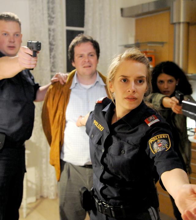 Das Bild zeigt eine spannungsgeladene Szene in einem Innenraum, vermutlich einer Wohnung oder einem Büro. Im Vordergrund stehen vier Personen. Eine Frau, gekleidet in eine Polizeiuniform, hält eine Waffe in der Hand und schaut entschlossen. Neben ihr steht ein Mann in einem hellen Hemd, der mit einem orangenen Mantel bedeckt ist; er hat eine besorgte Miene. Hinter ihm befinden sich zwei weitere Personen: eine Frau in einem olivfarbenen Outfit, die ebenfalls eine Waffe hält, und ein Mann in einer Polizeiuniform, der eine Waffe in Richtung des Betrachters richtet.  Der Raum hat helle Wände und eine moderne Holz-Küche im Hintergrund, die einige Küchengeräte und Ablagen zeigt. Vor den Fenstern befinden sich helle Vorhänge, die Licht hereinlassen. Die Pose der Charaktere deutet auf eine kritische Situation hin, in der sich alle Beteiligten in Alarmbereitschaft befinden.