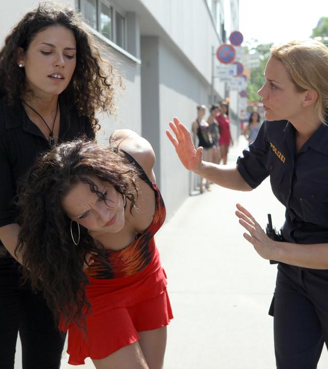 Das Bild zeigt eine Szene aus der Fernsehsendung "CopStories". Im Vordergrund sind drei Frauen abgebildet. Links steht eine Frau mit lockigem, dunklem Haar, die eine schwarze Bluse trägt und eine andere Frau festhält. Diese sieht erschöpft und verletzt aus, trägt ein rotes, ärmelloses Kleid und hat lange, dunkle, lockige Haare. Rechts von ihr steht eine Polizeibeamtin in einer blauen Uniform, die ein Symbol auf ihrer Schulter trägt, und hat eine betonte Körpersprache, als würde sie die Situation deeskalieren. Im Hintergrund sind weitere Personen und Gebäude zu sehen, typisch für eine städtische Umgebung, mit geparkten Autos auf der Straße. Die Atmosphäre wirkt angespannt.