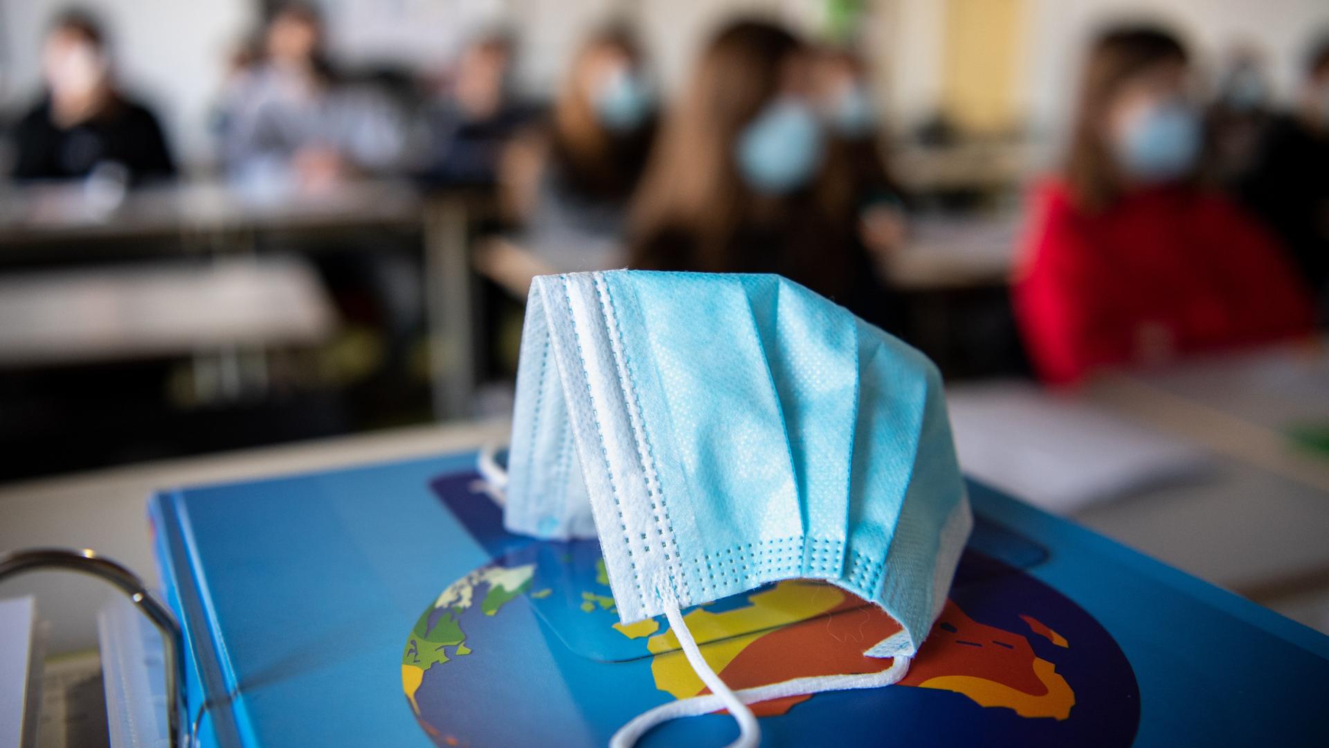 Ein Mund- und Nasenschutz liegt im Unterricht in einem Geographie-Seminar in der Jahrgangsstufe elf am staatlichen Gymnasium Trudering auf einem Weltalas, während im Hintergrund die Schülerinnen und Schüler mit Mund- und Nasenschutz zu sehen sind. 
