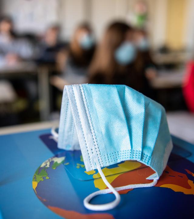 Ein Mund- und Nasenschutz liegt im Unterricht in einem Geographie-Seminar in der Jahrgangsstufe elf am staatlichen Gymnasium Trudering auf einem Weltalas, während im Hintergrund die Schülerinnen und Schüler mit Mund- und Nasenschutz zu sehen sind. 