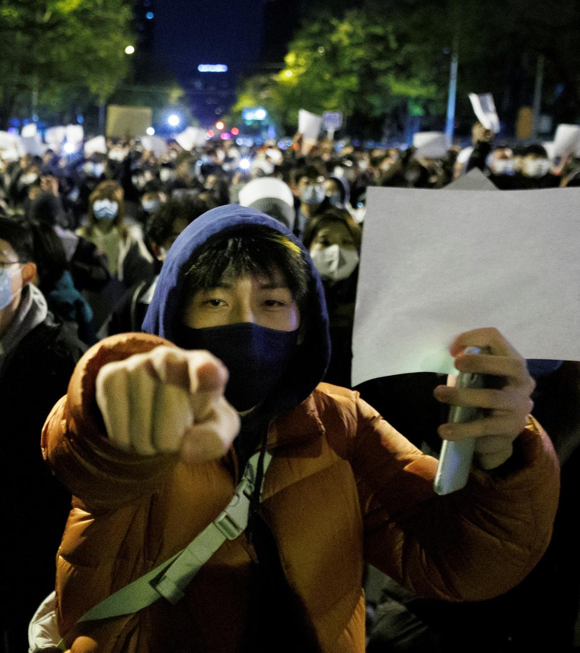 Eine Person mit Mundschutz hält ein leeres, weißes Papier in die Höhe. Im Hintergrund sieht man andere Demonstranten, die auch ein weißes Papier hochhalten.