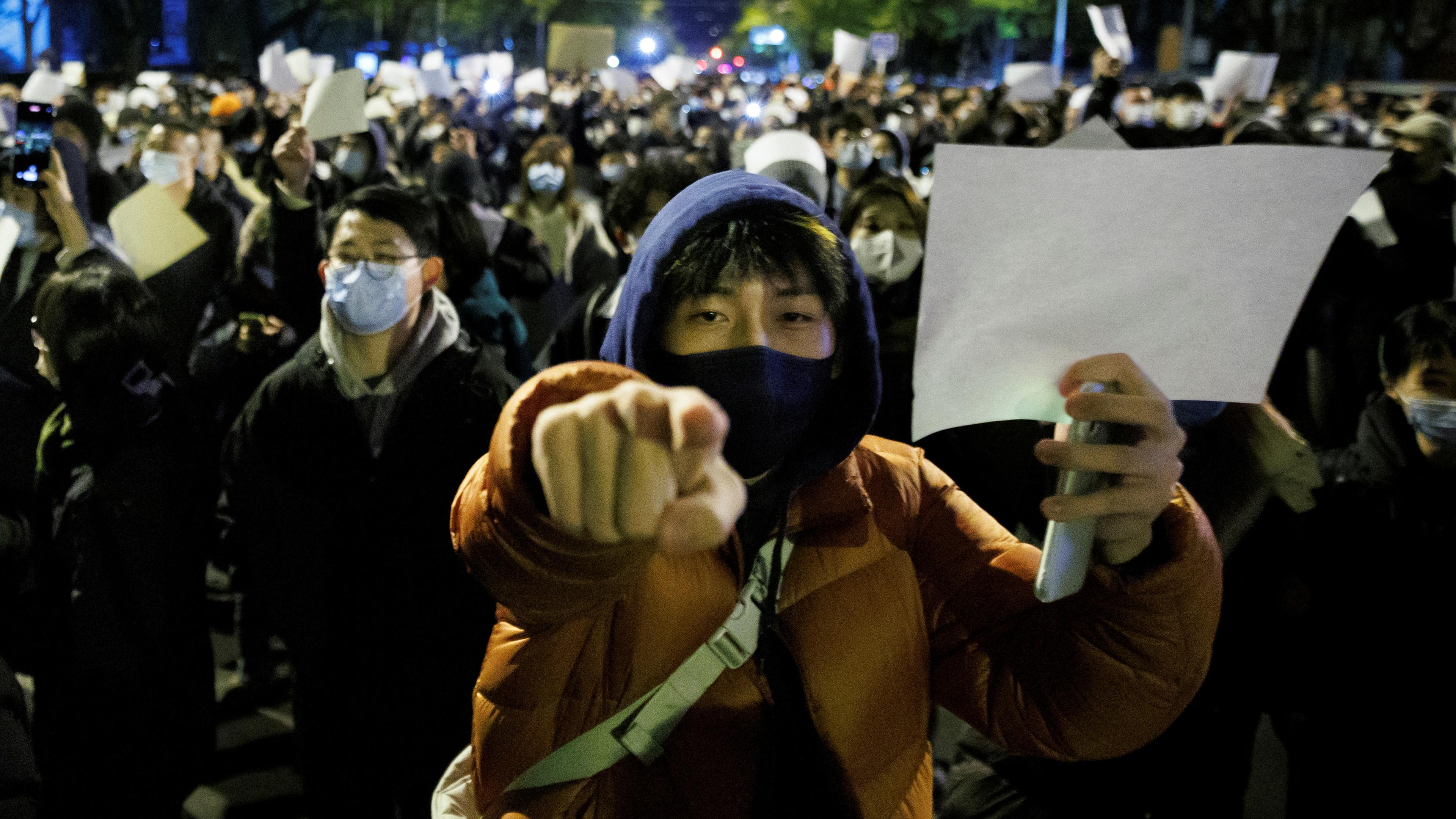 Eine Person mit Mundschutz hält ein leeres, weißes Papier in die Höhe. Im Hintergrund sieht man andere Demonstranten, die auch ein weißes Papier hochhalten.