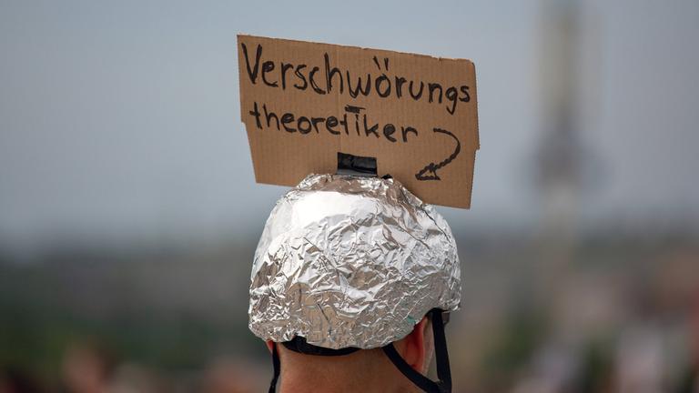 Ein Mann trägt einen Aluhut, an dem ein Schild mit der Aufschrift "Verschwörungstheoretiker" angebracht ist.