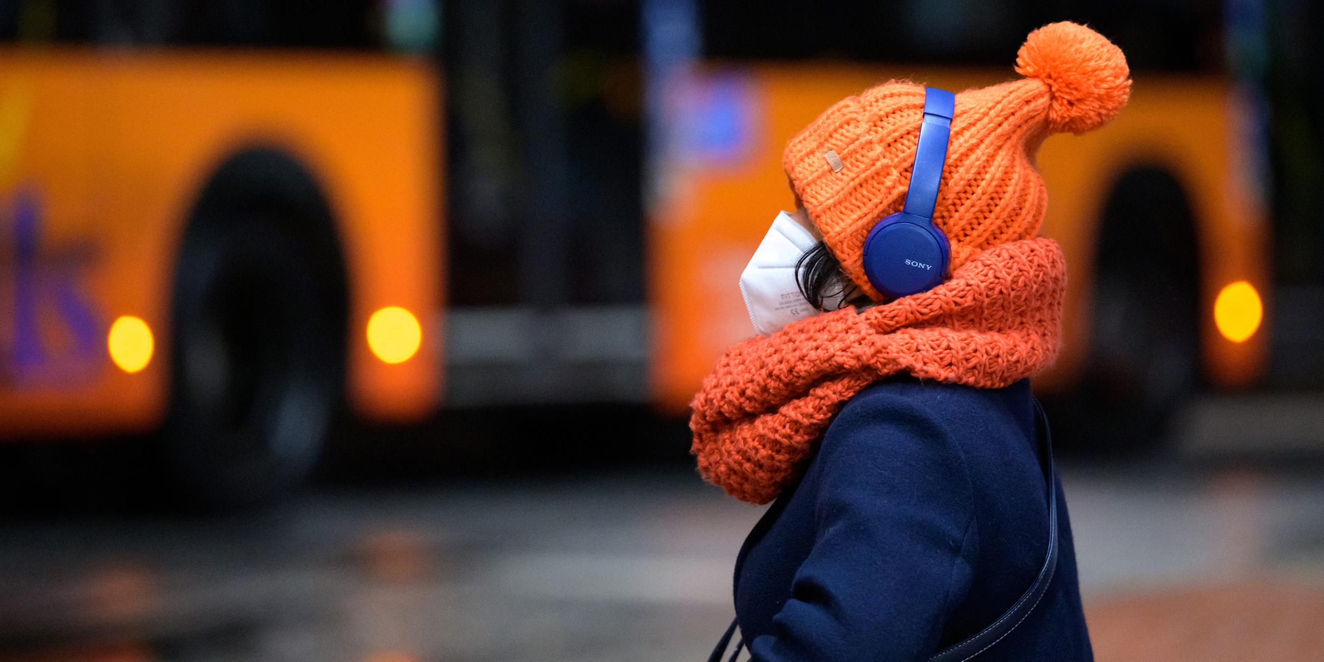 Eine Frau sitzt mit FFP2-Maske, dicker Mütze und Kopfhörern an einem Busbahnhof