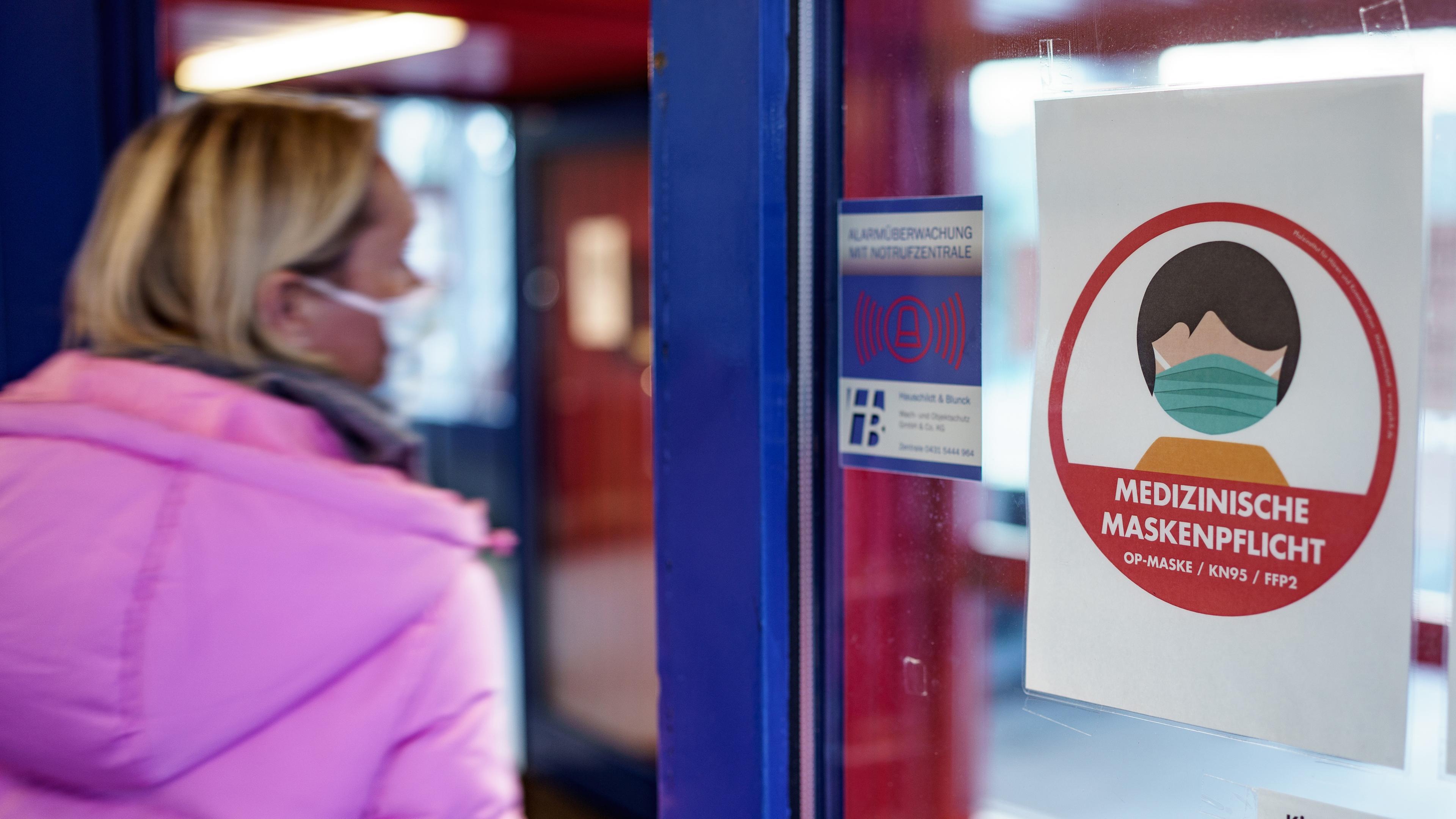 Schleswig-Holstein, Kiel: Eine Frau mit einer medizinischen Maske betritt ein Schwimmbad im Stadtteil Schilksee an dessen Eingang auf die Maskenpflicht hingewiesen wird.