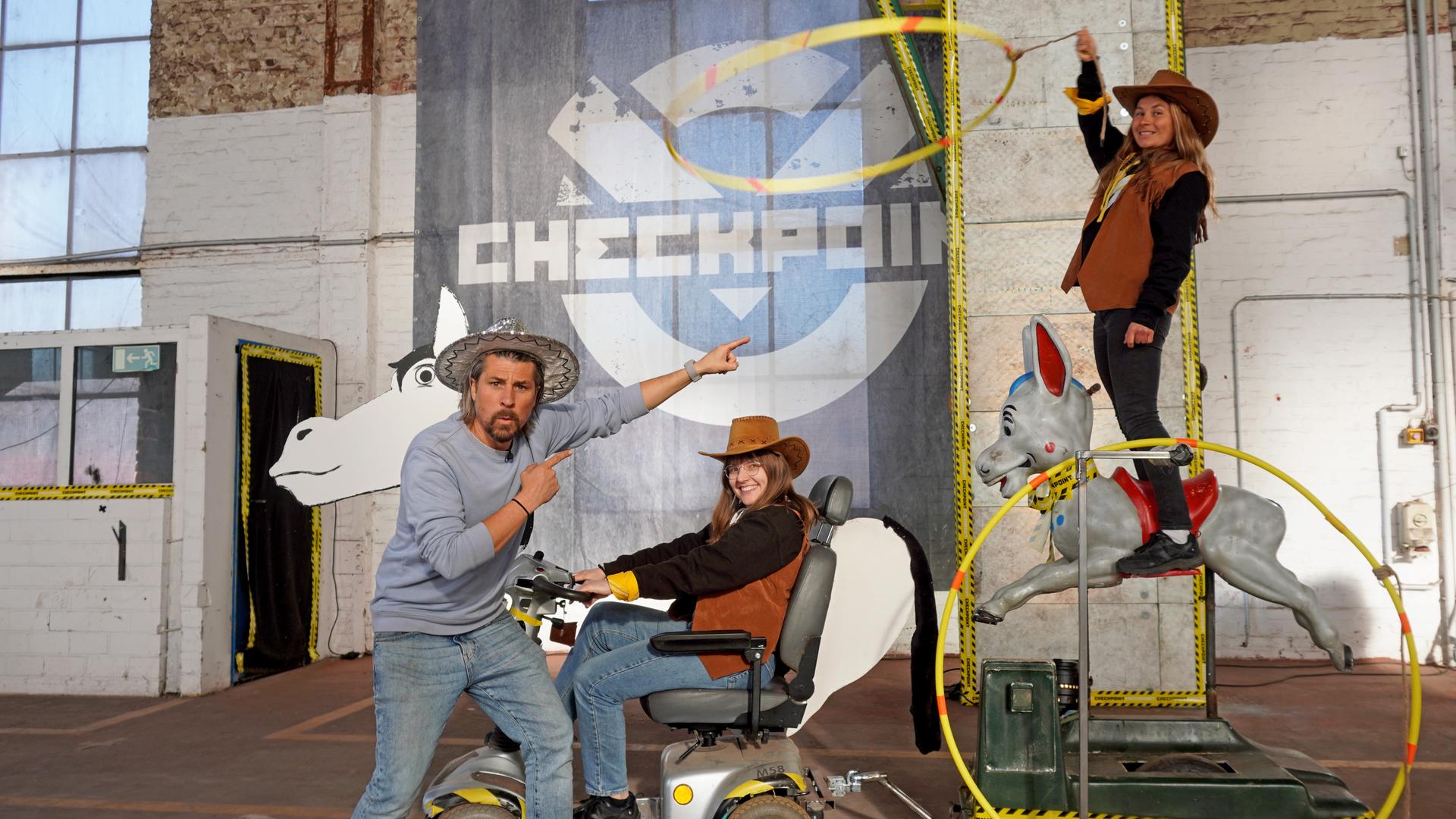 Checkpoint: Tommy Scheel zeigt auf Maja, die Lassoschwingend auf einem Karussell-Esel steht, der von einem Senioren-Scooter gezogen wird, der von Lisa gefahren wird. 
